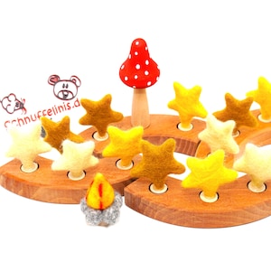 Könnte beinhalten: Holzspielzeug mit einem roten Pilz und sternförmigen Filzstücken in Gelb- und Brauntönen. Das Spielzeug hat eine Holzbasis mit Löchern für die Sterne und ein kleines Filz-Lagerfeuer. Das Logo "Schnuffelinis.d" ist sichtbar.