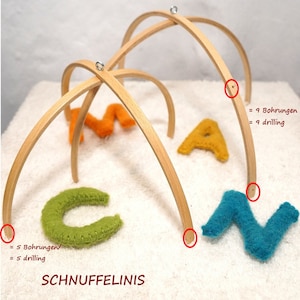 Può includere: Un telaio per mobile in legno con nove fori praticati. Il telaio è in legno naturale ed è progettato per contenere lettere in feltro. Le lettere sono di colore arancione, giallo, verde e blu. Il testo "SCHNUFFELINIS" è stampato sulla superficie bianca sotto il telaio.