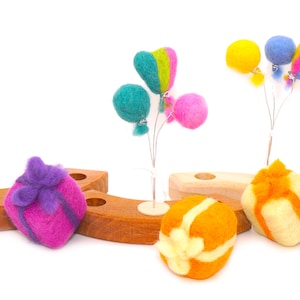 Könnte beinhalten: Eine Sammlung farbenfroher Filzdekorationen, darunter Geschenkboxen mit lila Schleifen, orange-weiß gestreifte Kugeln und Ballongruppen in verschiedenen Farben. Die Ballons sind türkis, rosa, gelb und blau. Die Dekorationen befinden sich auf Holzblöcken.