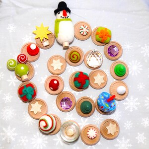 May include: A collection of decorative wooden discs and ornaments, including a snowman figure, star shapes, and various colorful spheres and gems. The ornaments are arranged on a white surface with snowflake patterns.