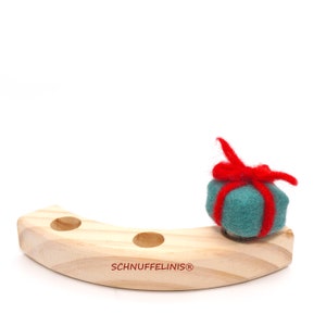 May include: A wooden, crescent-shaped stand with three holes, featuring the text "SCHNUFFELINIS&reg;". A teal felt gift box with a red felt bow sits on the stand.