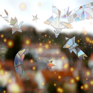 Könnte beinhalten: Ein Fenster mit Blick auf eine verschwommene Stadtlandschaft und Lichter. Das Fenster ist mit schimmernden Sternen- und Halbmondaufklebern verziert.