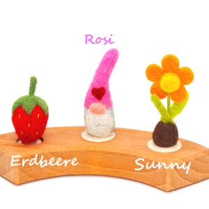 Puede incluir: Una exhibición de madera con tres figuras de fieltro: una fresa roja, un gnomo rosa con un corazón y una flor naranja. La fresa tiene un tallo verde, el gnomo tiene barba blanca y la flor tiene un centro amarillo. La palabra "Rosi" está escrita encima del gnomo.
