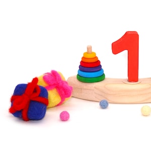 Könnte beinhalten: Ein Holzspielzeug-Set mit einer roten Zahl 1, einem bunten Stapelring-Spielzeug und zwei Filz-Geschenkboxen mit Schleifen. Kleine Filzbälle in Blau, Rosa und Gelb sind um die Spielzeuge herum verstreut.