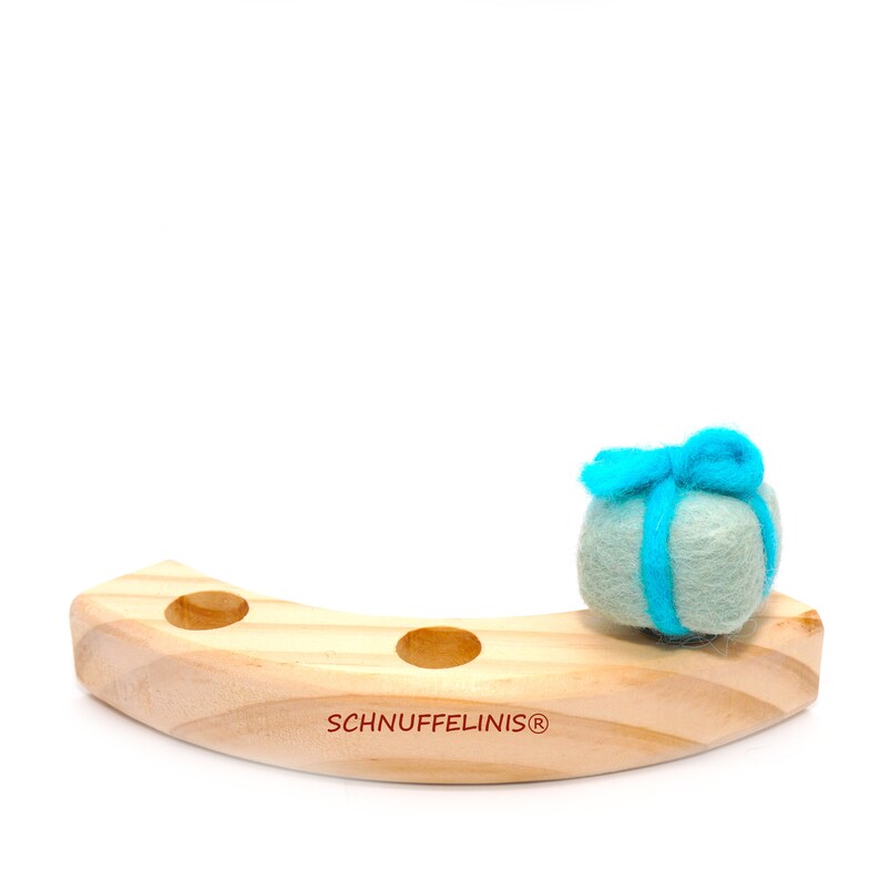 May include: A wooden, curved stand with three holes, featuring the text "SCHNUFFELINIS&reg;" on the bottom. A light blue felt ball with a blue ribbon is sitting on the stand.