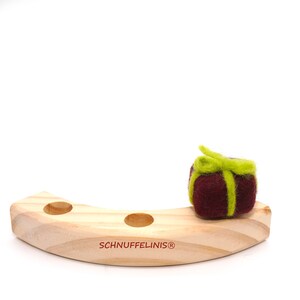 May include: A wooden toy with three holes and a felt gift with a green bow on top. The toy is labeled "Schnufelinis&reg;".