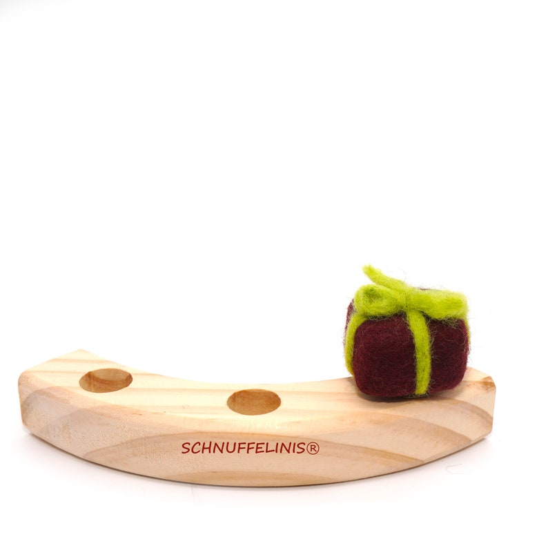 May include: A wooden toy with three holes and a felt gift with a green bow on top. The toy is labeled "Schnufelinis&reg;".
