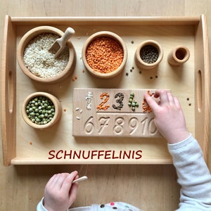 Op de afbeelding: Een houten dienblad met kleine houten kommen gevuld met rijst, linzen en erwten. Een houten cijferbord met de cijfers 1-10 is gedeeltelijk gevuld met granen en peulvruchten. Het woord "SCHNUFFELINIS" is in het rood geschreven.