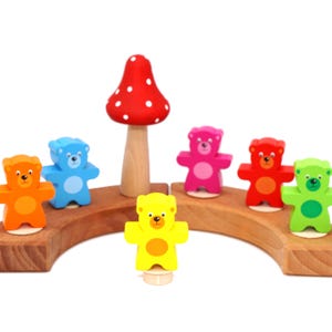 Könnte beinhalten: Ein Holzspielzeug-Set mit bunten Bärenfiguren in Orange, Blau, Pink, Rot, Gelb und Grün sowie einem roten Pilz mit weißen Punkten. Die Bären sind auf einem Holz-Bogen angeordnet.
