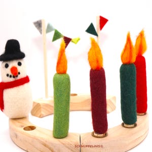 May include: A needle-felted snowman with a black hat and red scarf stands on a curved wooden base. Beside it are four felt candles in green, maroon, dark green, and red, each with an orange flame, plus small felt pennant flags. Text reads SCHNUFFELINIS.