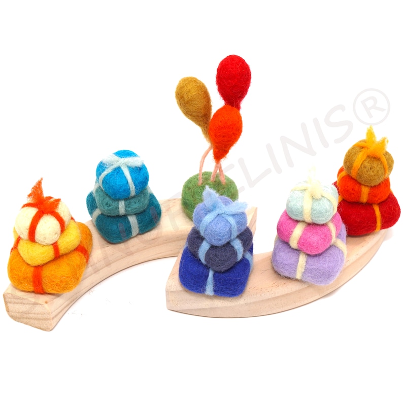 Op de afbeelding: Een houten regenboogvormige basis met vijf stapels viltgeschenken in verschillende kleuren, waaronder geel, oranje, rood, roze, paars, blauw, groen en teal. Een groene viltbasis met twee rode en oranje ballonnen staat op de basis.