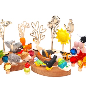 Op de afbeelding: Een verzameling houten dierenfiguren waaronder een vleermuis, eekhoorn, egel, gans, cavia en vogel, samen met viltbloemen, een zon en doorschijnende gekleurde edelstenen. De set bevat lasergesneden houten plantensilhouetten op houten voetjes.