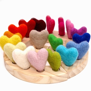 Op de afbeelding: Een verzameling kleine hartjes van viltwol in een spectrum van kleuren, waaronder rood, oranje, geel, beige, roze, groen, blauw en paars. Elk hartje is afzonderlijk bevestigd in een ronde, lichtgekleurde houten basis met geboorde gaten.