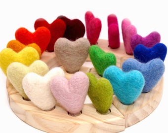May include: A collection of small, felted wool hearts in a spectrum of colors including red, orange, yellow, beige, pink, green, blue, and purple. Each heart is individually mounted in a circular, light-colored wooden base with drilled holes.