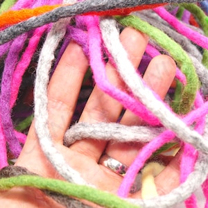 May include: Close-up of colorful felted wool roving in shades of pink, purple, green, gray, and orange. The wool is tightly coiled and held in a hand, showcasing the texture and variety of colors. Ideal for fiber art projects.