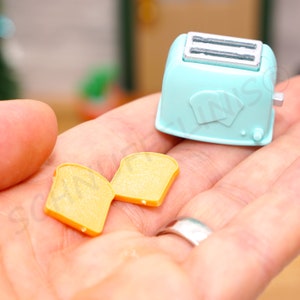 Op de afbeelding: Een miniatuur turquoise broodrooster met een zilveren bovenkant en twee sneetjes toast. De broodrooster heeft een eenvoudig ontwerp met twee toastvormige uitsparingen. De toastjes zijn geel met oranje randen.