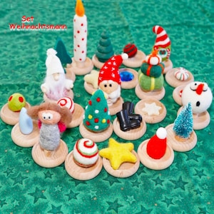 May include: A collection of Christmas-themed felt ornaments on wooden bases. The set includes miniature trees, gnomes, a candle, a star, and other festive shapes. The text "Set Weihnachtsmann" is visible in the upper left corner. The ornaments are arranged on a green surface.
