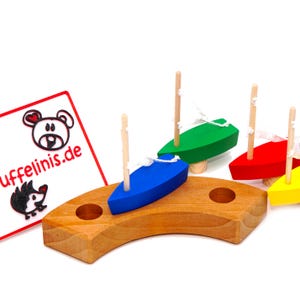 Könnte beinhalten: Holzspielzeugboote in Blau, Grün, Rot und Gelb mit weißen Segeln und Holzmasten auf einer gebogenen Holzbasis. Eine weiße Karte mit dem Text "uffelinis.de" und einem Bären- und Igel-Logo ist links zu sehen.