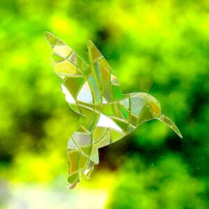 May include: A stained glass hummingbird silhouette in shades of green, yellow, and white. The bird's form is composed of irregular geometric shapes, creating a mosaic effect. The background is a blurred, vibrant green.