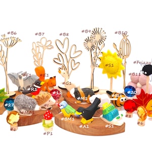Op de afbeelding: Een verzameling kleine houten dierenfiguren, viltvormen en decoratieve plantenuitsparingen gelabeld met alfanumerieke codes. De set bevat een vleermuis, eekhoorn, gans, egel, vogel en slak, naast kleurrijke glazen edelstenen en een gele zon.