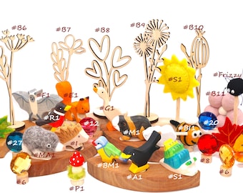 May include: A collection of small wooden animal figurines, felt shapes, and decorative plant cutouts labeled with alphanumeric codes. The set includes a bat, squirrel, goose, hedgehog, bird, and snail, alongside colorful glass gems and a yellow sun.