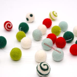 Könnte beinhalten: Eine Sammlung bunter Filzkugeln in verschiedenen Größen und Farben, darunter Rot, Grün, Weiß und Hellblau. Einige Kugeln haben Spiral- oder Tupfenmuster. Der Text "SCHNUFFELINIS®" ist auf einer der Kugeln aufgedruckt. Die Kugeln sind aufgefädelt.