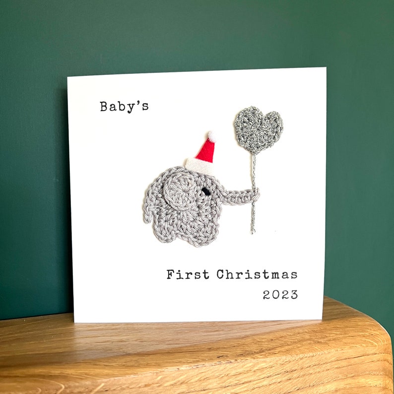 May include: A white card with the text "Baby's First Christmas 2023". It features a crocheted grey elephant wearing a red and white Santa hat, holding a grey heart-shaped balloon.