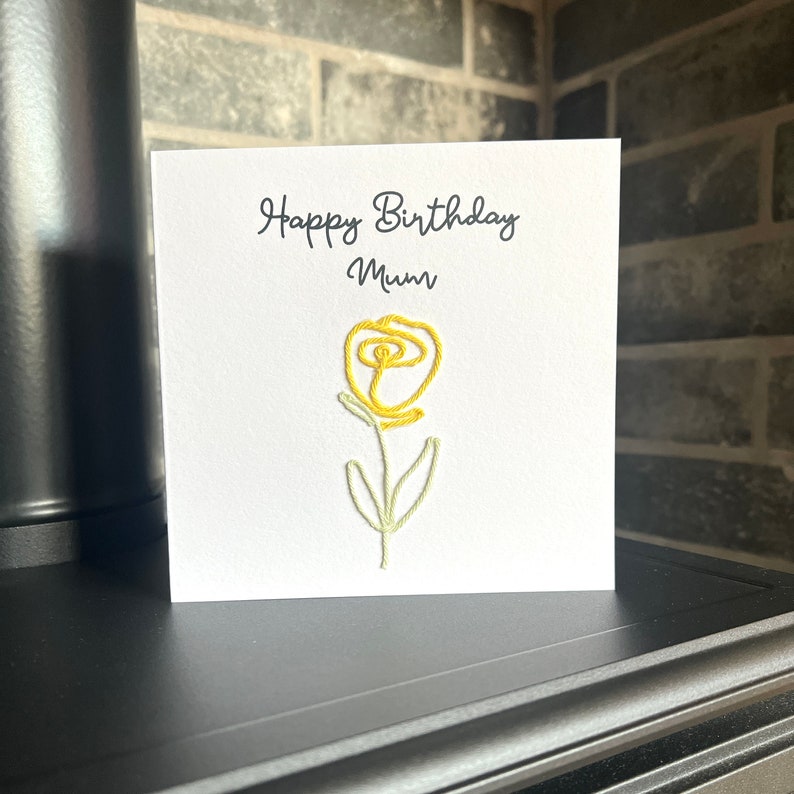Peut inclure: Une carte blanche avec le texte "Happy Birthday Mum" et une fleur brod&eacute;e jaune.