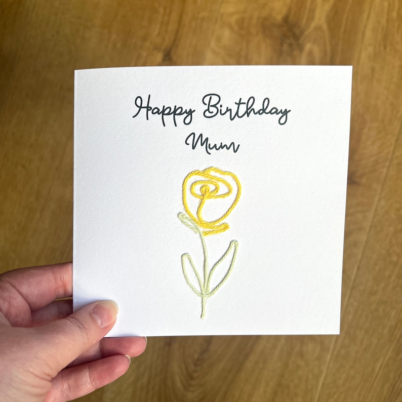 Peut inclure: Une carte blanche avec le texte "Happy Birthday Mum" et une fleur brod&eacute;e jaune.