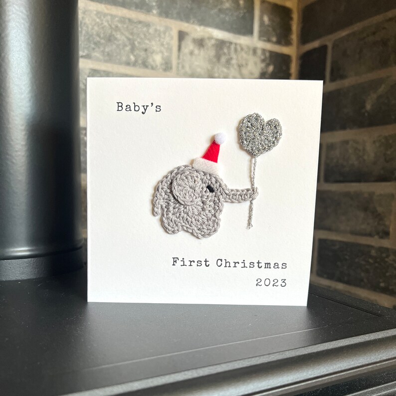 May include: A handmade card featuring a crocheted grey elephant wearing a red Santa hat and holding a grey heart-shaped balloon. The card reads "Baby's First Christmas 2023" in black text on a white background.