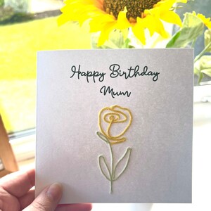 Peut inclure: Une carte blanche avec le texte "Happy Birthday Mum" et une fleur brod&eacute;e jaune.