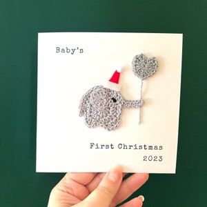 May include: A handmade Christmas card with a crocheted grey elephant wearing a Santa hat and holding a heart-shaped balloon. The text reads "Baby's First Christmas 2023" in black font on a white card.