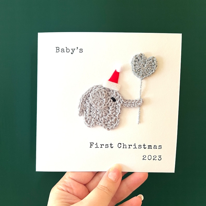 May include: A handmade Christmas card with a crocheted grey elephant wearing a Santa hat and holding a heart-shaped balloon. The text reads "Baby's First Christmas 2023" in black font on a white card.