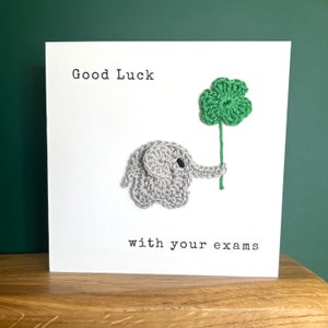 May include: A white card with the text "Good Luck with your exams" and a crocheted gray elephant holding a green four-leaf clover.