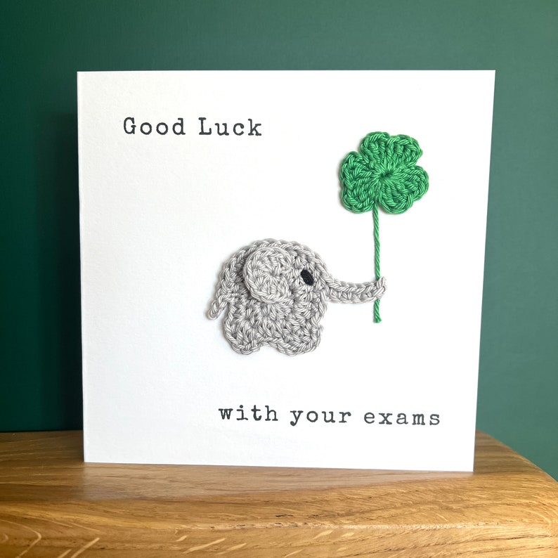 May include: A white card with the text "Good Luck with your exams" and a crocheted gray elephant holding a green four-leaf clover.