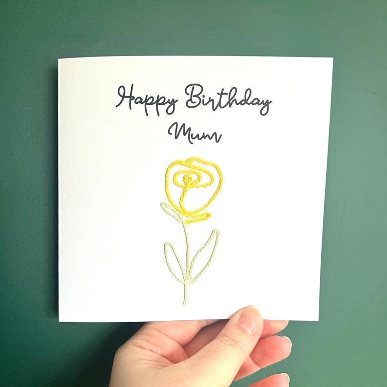 Peut inclure: Une carte blanche avec le texte "Happy Birthday Mum" et une fleur brod&eacute;e jaune.