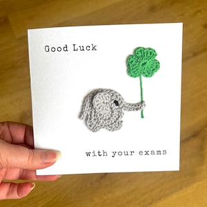 May include: A white card with the text "Good Luck with your exams" and a crocheted gray elephant holding a green four-leaf clover.