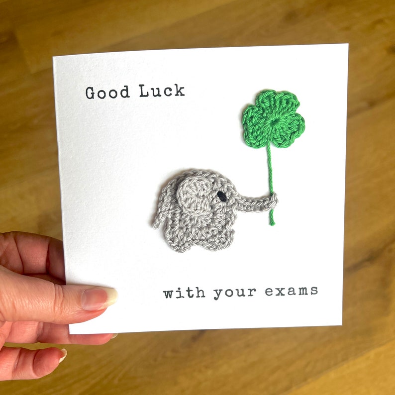 May include: A white card with the text "Good Luck with your exams" and a crocheted gray elephant holding a green four-leaf clover.