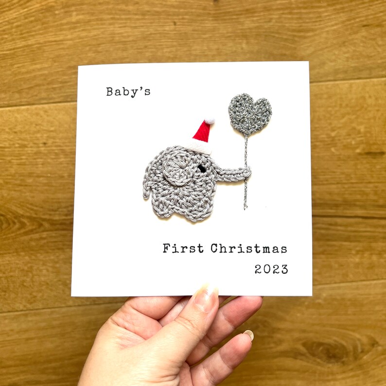 May include: A white card with the text "Baby's First Christmas 2023". It features a crocheted grey elephant wearing a red Santa hat and holding a silver heart-shaped balloon.