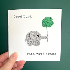 May include: A white card with the text "Good Luck with your exams" and a crocheted gray elephant holding a green four-leaf clover.
