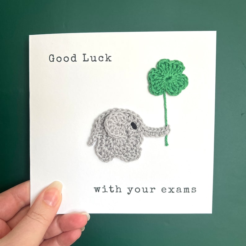 May include: A white card with the text "Good Luck with your exams" and a crocheted gray elephant holding a green four-leaf clover.