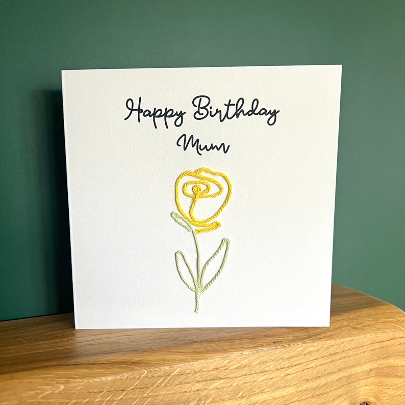 Peut inclure: Une carte blanche avec le texte "Happy Birthday Mum" et une fleur brod&eacute;e jaune.