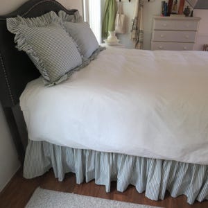 May include: A bed with a dark gray upholstered headboard, white bedding, and striped pillows with ruffled edges. The bed skirt is also striped in the same pattern. The room has a neutral color palette.