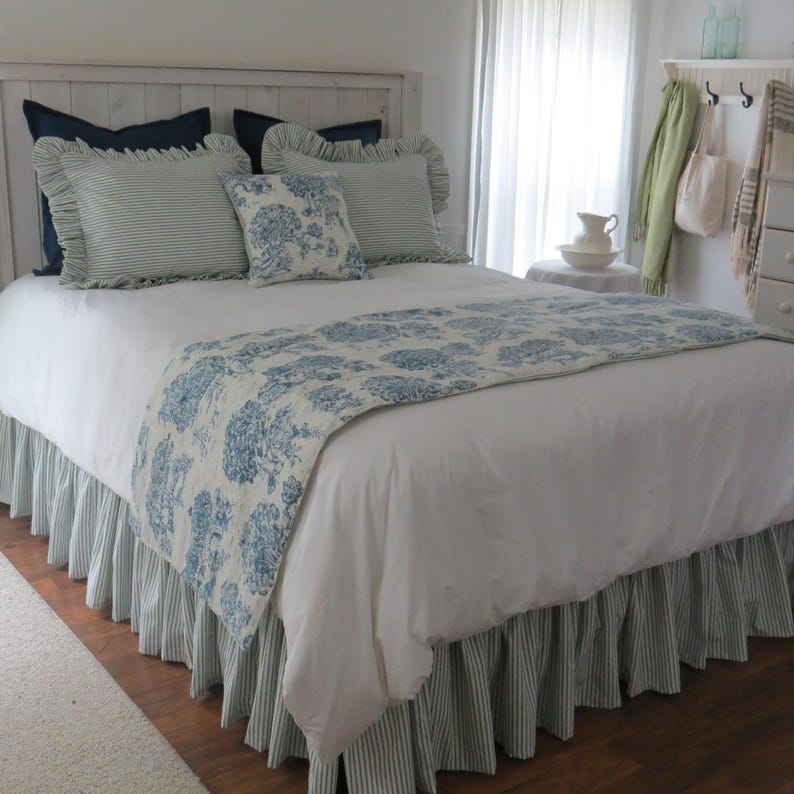 May include: A bed with white bedding, a blue floral runner, and striped bed skirt. Pillows include solid navy, striped, and floral patterns. The headboard is white, and the room has a neutral color palette.
