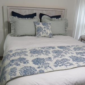 May include: A bed with a white comforter and a blue and white patterned bed runner. The bed has several pillows, including striped and solid blue pillows, and a decorative pillow with a tree design. The headboard is made of light-colored wood.