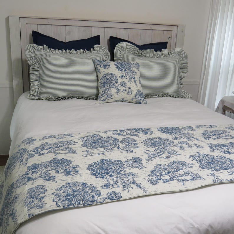 May include: A bed with a white comforter and a blue and white patterned bed runner. The bed has several pillows, including striped and solid blue pillows, and a decorative pillow with a tree design. The headboard is made of light-colored wood.