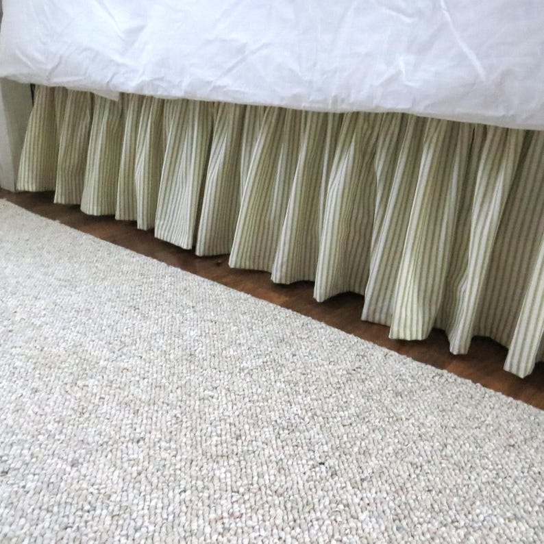 May include: A white bed with a green and white striped bed skirt. The bed skirt is pleated and has a ruffled edge. The bed skirt is on a wooden floor.