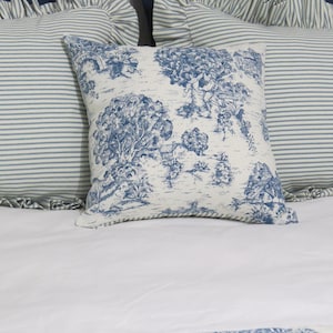 May include: Square throw pillow with a blue and white toile design featuring a landscape scene. The pillow is placed on a white bed with two striped pillows in the background. The pillow has a soft, textured appearance.