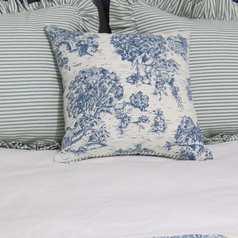 May include: Square throw pillow with a blue and white toile design featuring a landscape scene. The pillow is placed on a white bed with two striped pillows in the background. The pillow has a soft, textured appearance.