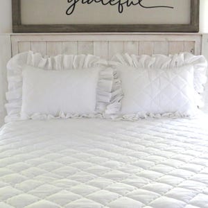 May include: White ruffled pillowcases on a white quilted bedspread. The headboard is white wood with a rustic finish.